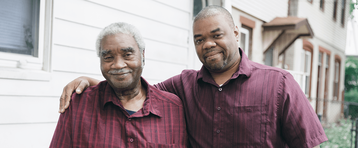 Two men stand outside next to a white house, both wearing maroon shirts. The younger man has his arm around the older mans shoulder. They are smiling and looking at the camera.