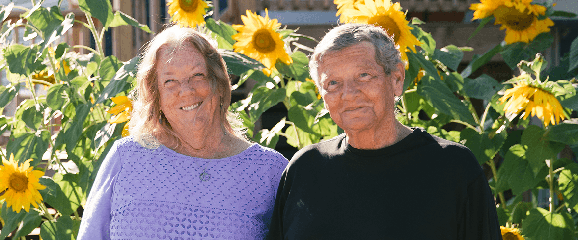 Two older adults, one in a lavender top and the other in a black shirt, stand smiling in front of tall sunflowers on a sunny day.